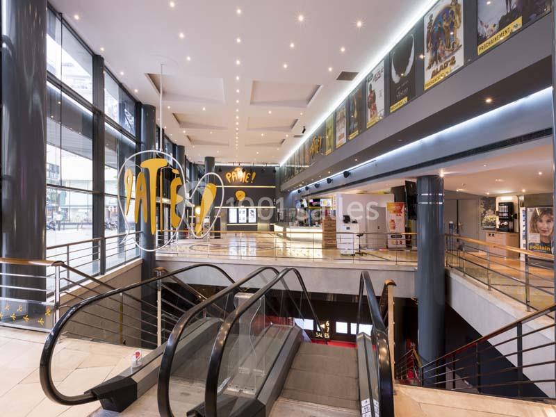 Intérieur d'un cinéma avec escalators, affiches de films sur les murs et guichet visible au fond.