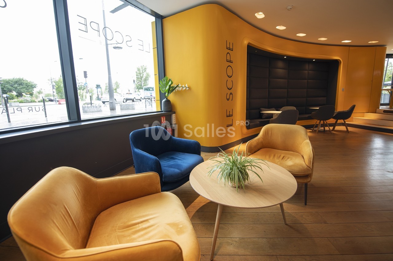 Espace d'attente moderne avec fauteuils colorés, table basse et murs jaunes, éclairé par de grandes fenêtres.
