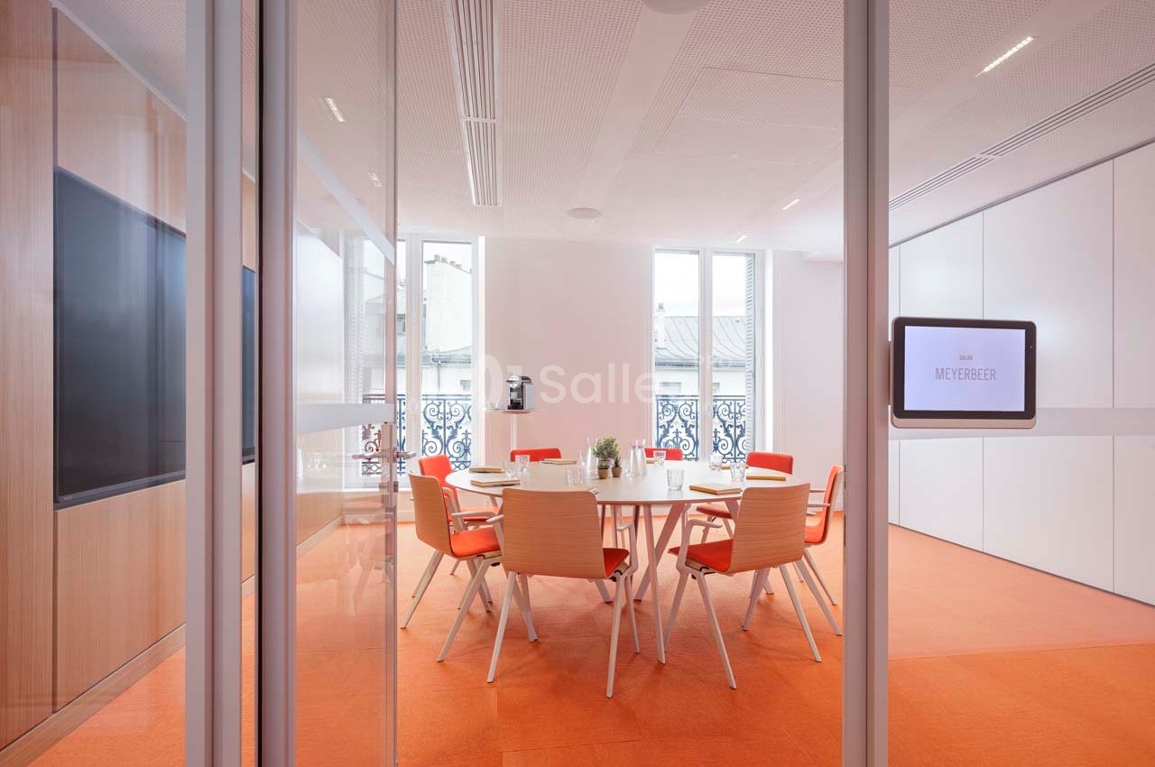 Salle de réunion lumineuse avec table ronde, chaises orange et vue sur des fenêtres donnant sur des balcons.