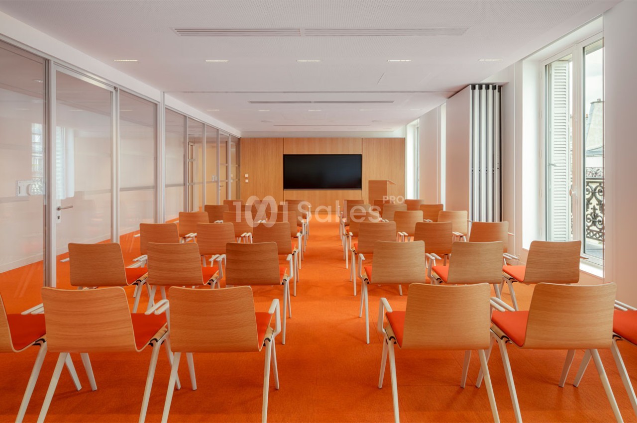 Salle de conférence lumineuse avec des rangées de chaises en bois et un écran noir fixé sur un mur en bois clair.