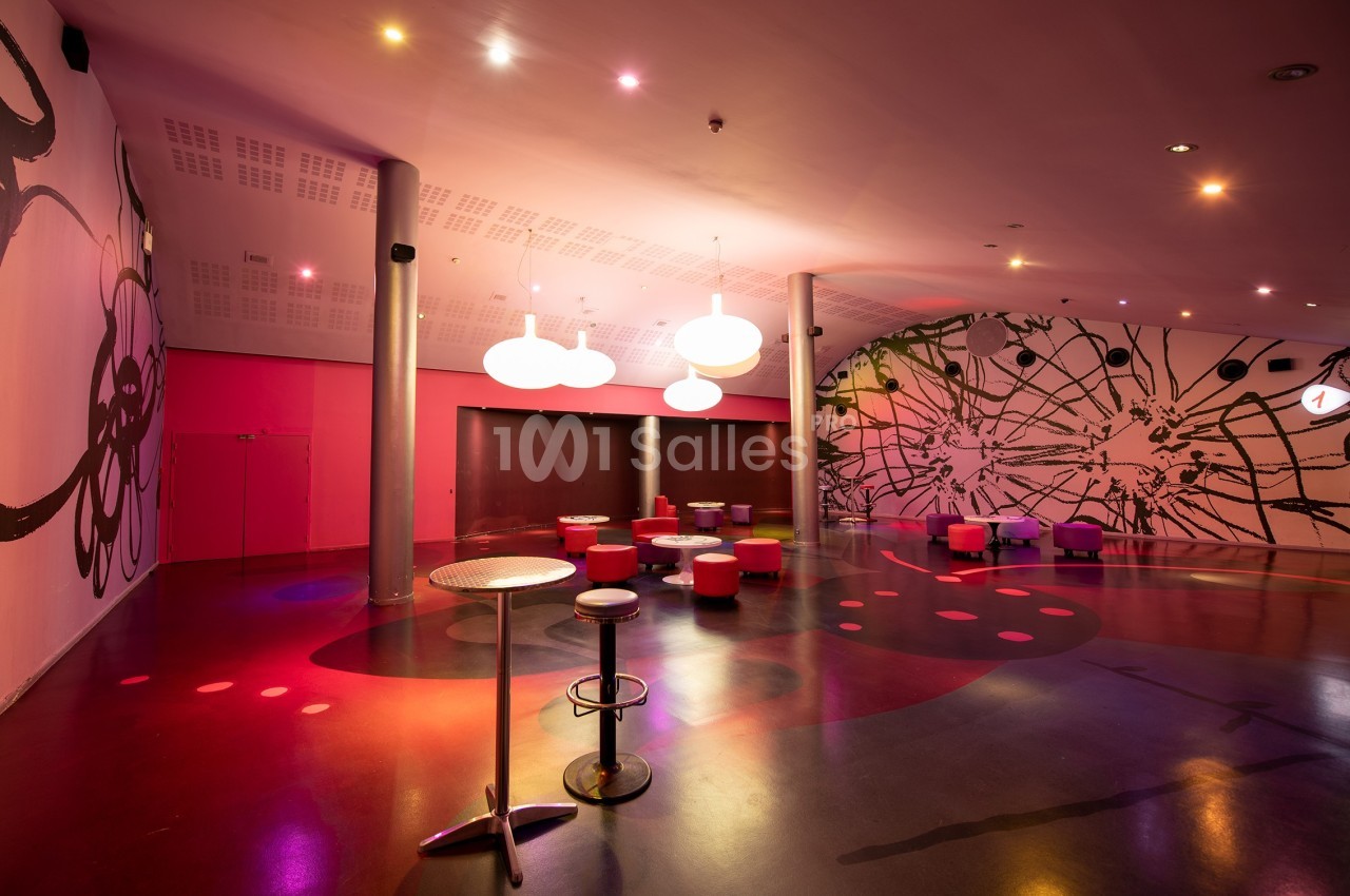 Salle moderne avec éclairage coloré, murs décorés de motifs abstraits, tables hautes et sièges dispersés.