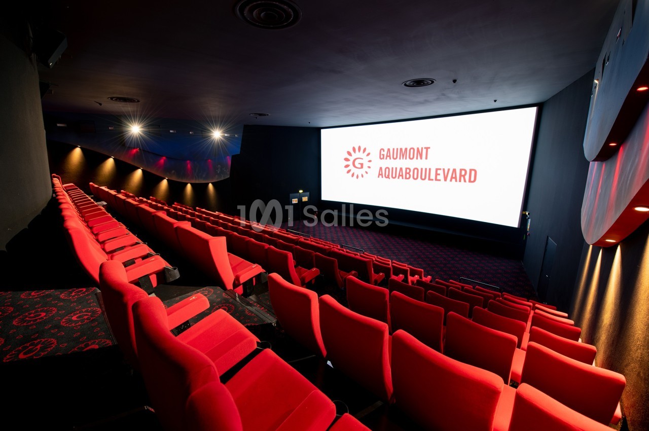 Salle de cinéma vide avec des fauteuils rouges, écran blanc affichant ’Gaumont Aquaboulevard’ et éclairage tamisé.