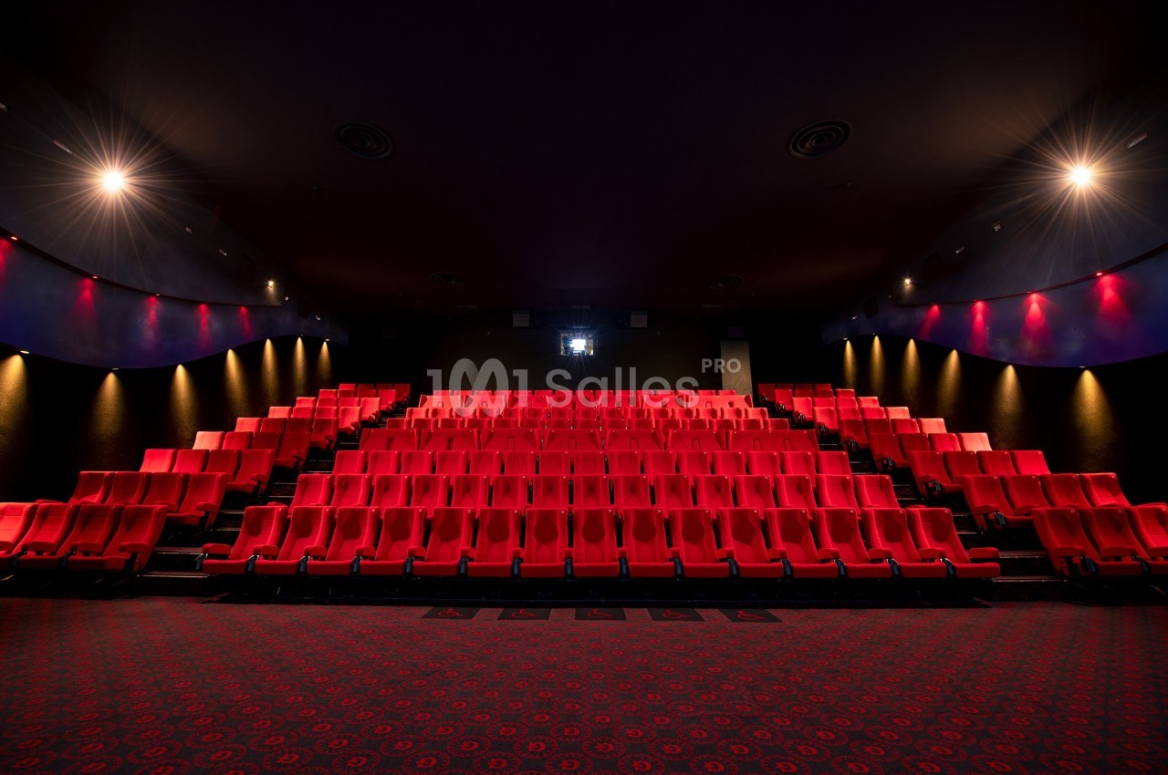 Salle de cinéma vide avec des rangées de sièges rouges et un écran blanc au fond, éclairée par des lumières tamisées.