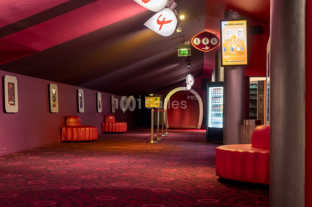 Couloir d'un cinéma avec moquette rouge, sièges arrondis, distributeurs automatiques et signalétique lumineuse.