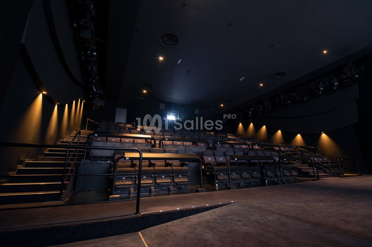 Salle de théâtre vide avec rangées de sièges, éclairage tamisé et scène au premier plan.