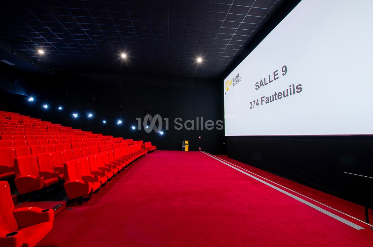 Salle de cinéma avec des sièges rouges, un écran géant affichant ’Salle 9, 374 fauteuils’ et un sol recouvert de moquette…