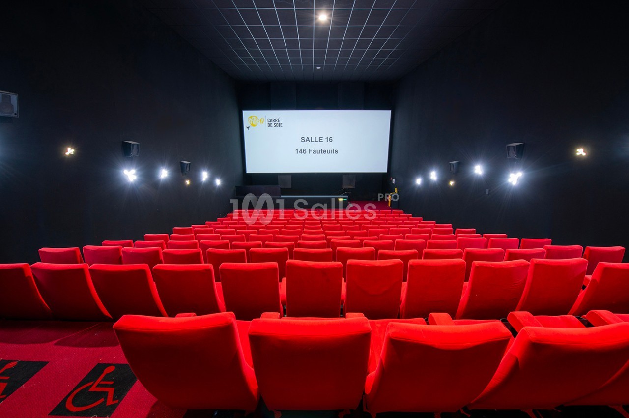 Salle de cinéma avec des rangées de fauteuils rouges vides, écran blanc et éclairage tamisé.