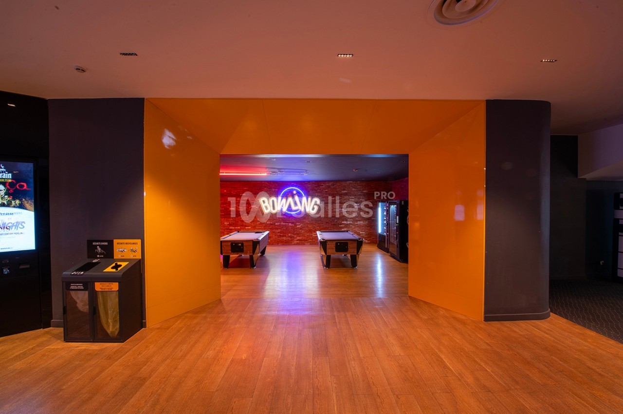 Entrée d'une salle de bowling avec un mur en briques, un néon ’Bowling’ et des tables de jeu en premier plan.