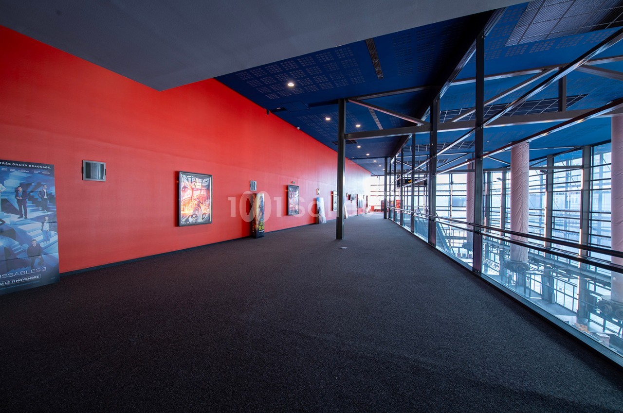 Couloir spacieux avec mur rouge, affiches encadrées, sol en moquette sombre et grandes baies vitrées à droite.