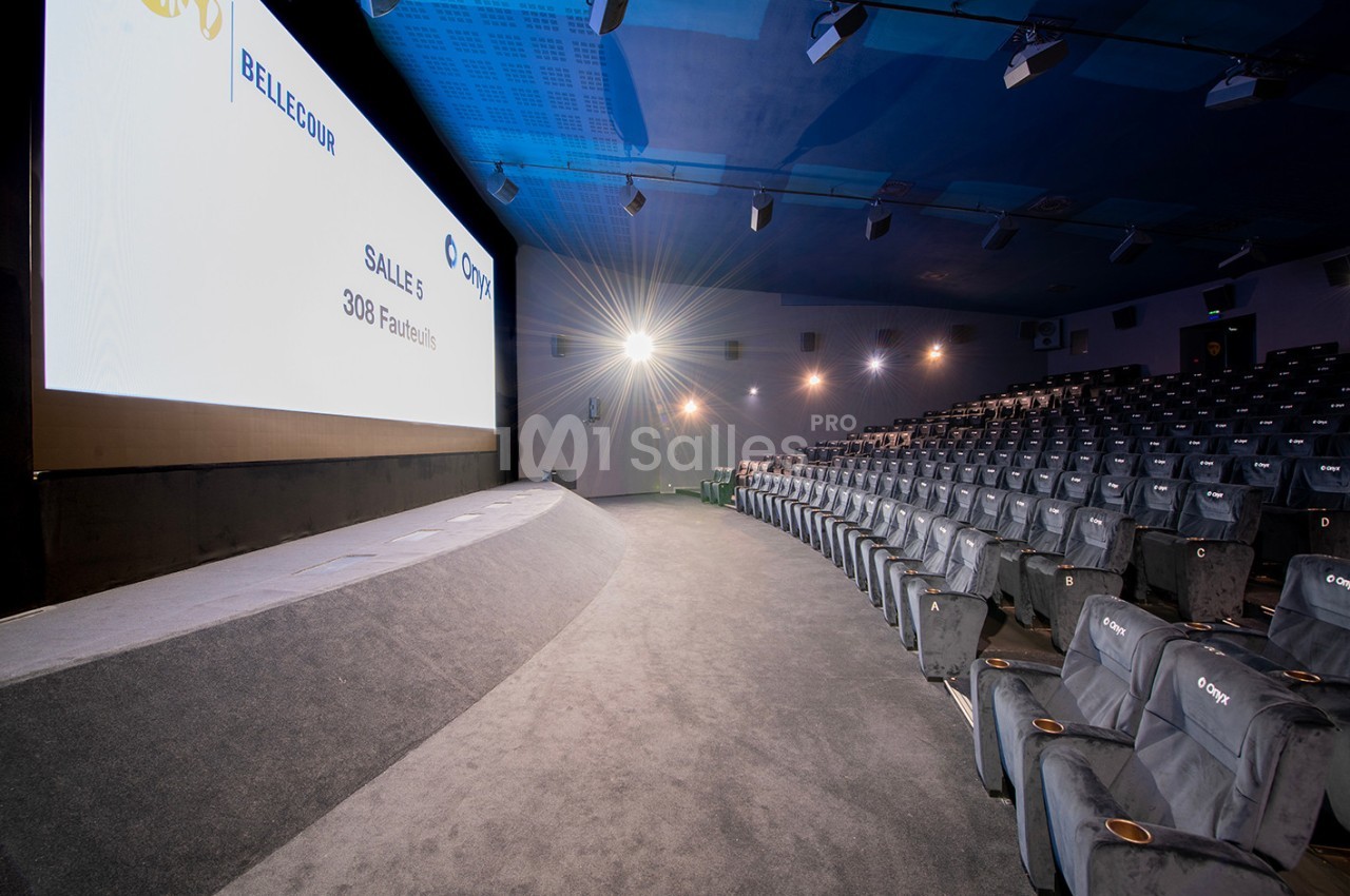 Salle de cinéma vide avec des rangées de sièges gris, un grand écran allumé et un éclairage tamisé.