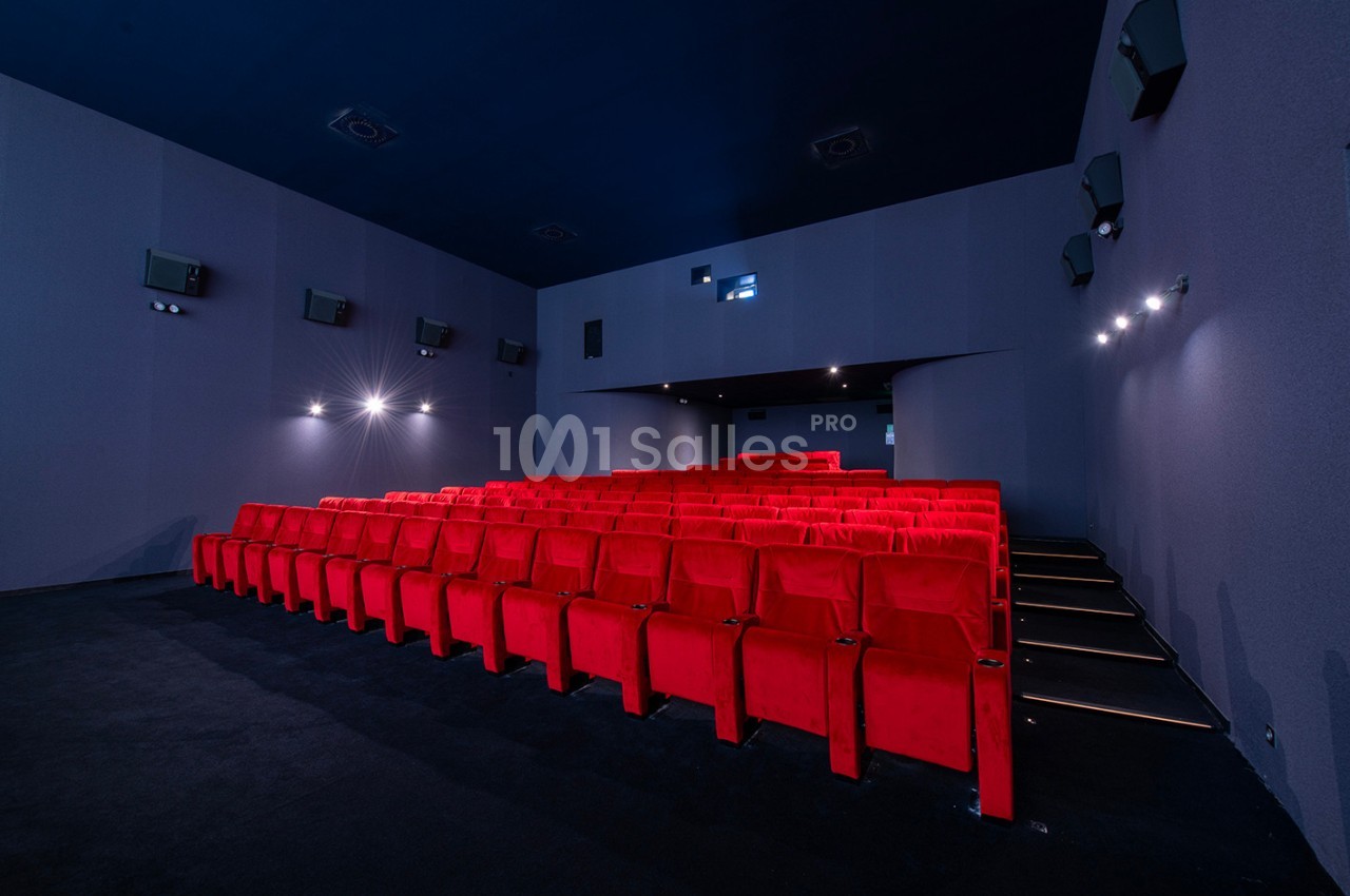 Salle de cinéma vide avec des rangées de fauteuils rouges, murs gris et éclairage tamisé.
