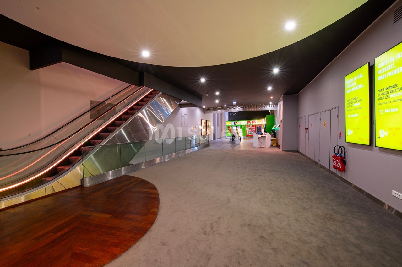Vue d'un espace intérieur moderne avec escalator, écrans d'affichage lumineux et comptoir d'accueil au fond.