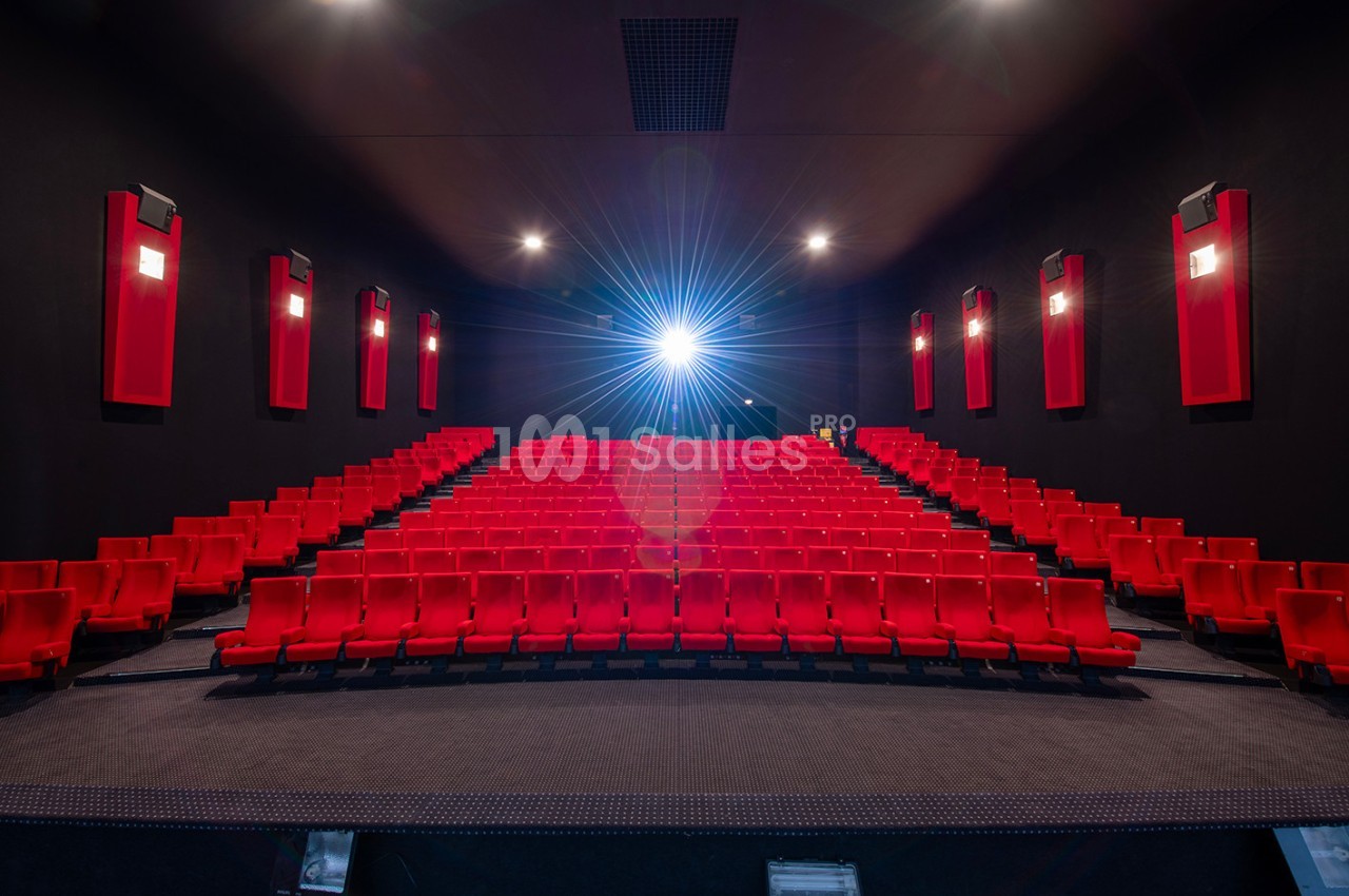 Salle de cinéma vide avec des rangées de sièges rouges et un projecteur allumé au fond.