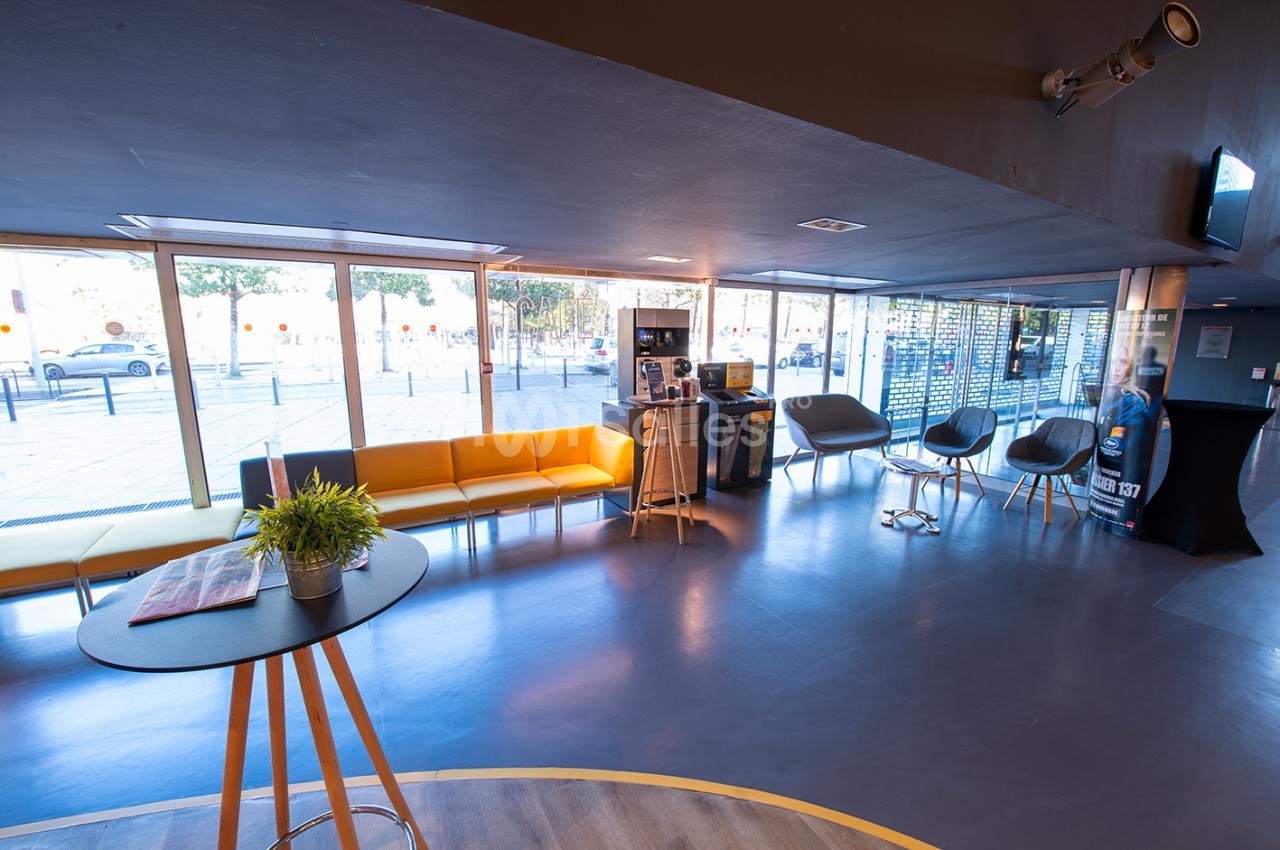 Espace d'accueil lumineux avec canapés jaunes, chaises grises, table haute et grandes baies vitrées donnant sur l'extérieur.