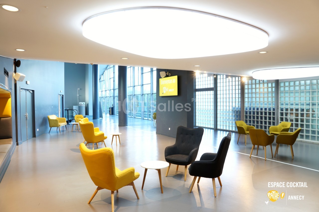 Salle lumineuse avec fauteuils colorés, tables basses et grande baie vitrée donnant sur l'extérieur.