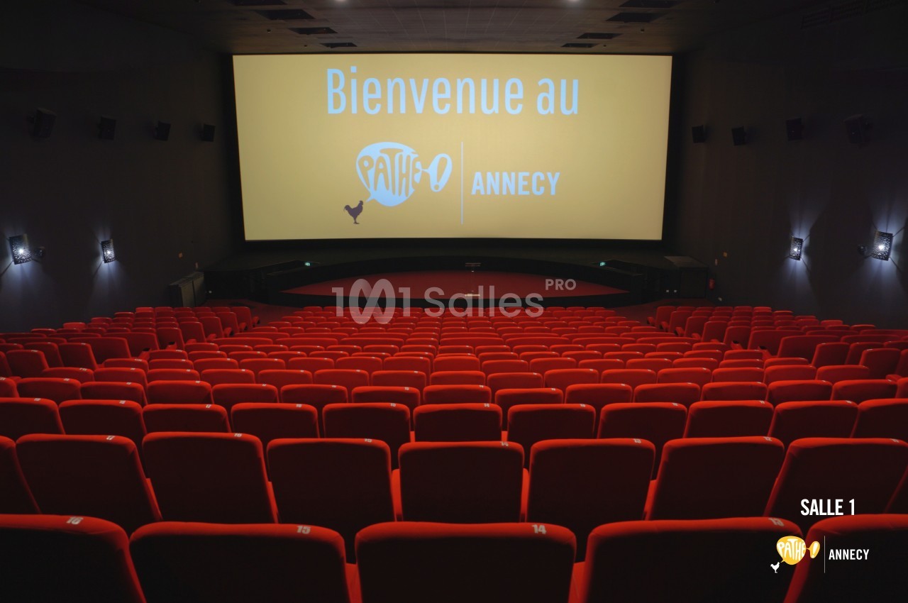 Salle de cinéma vide avec des rangées de sièges rouges et un écran affichant ’Bienvenue au Pathé Annecy’.
