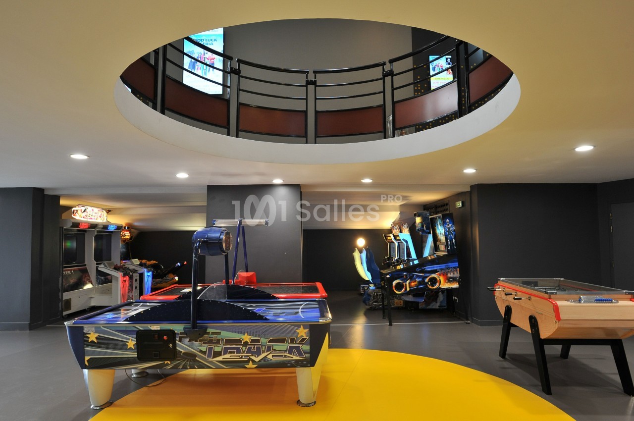 Salle de jeux avec un air hockey, un baby-foot et des bornes d'arcade sous un plafond circulaire ouvert.