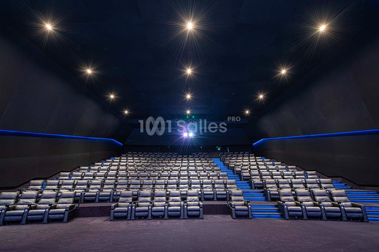 Salle de cinéma vide avec rangées de sièges inclinables, éclairage tamisé et projecteur allumé.