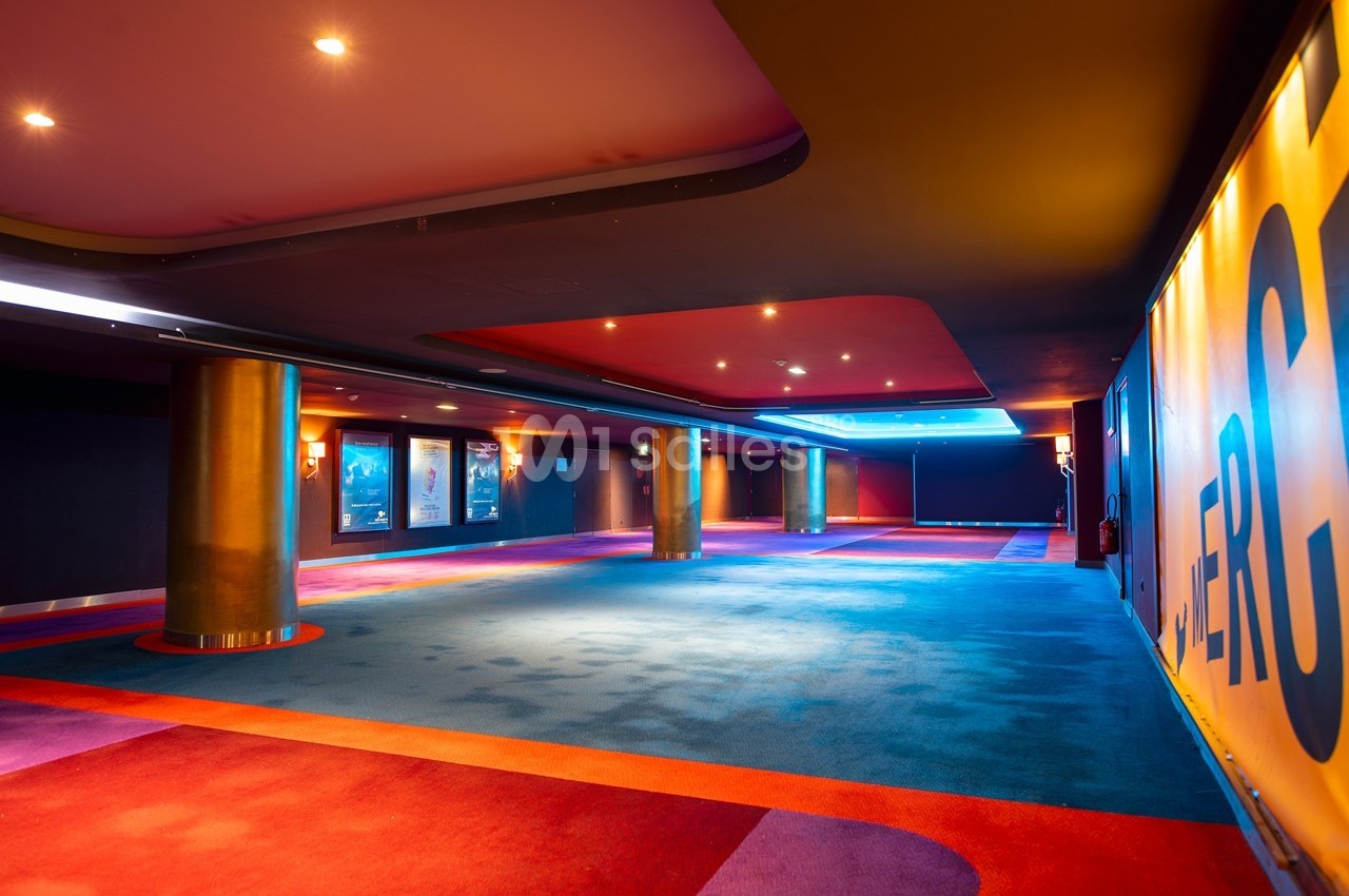 Couloir spacieux d'un cinéma avec moquette colorée, affiches de films aux murs et éclairage tamisé.