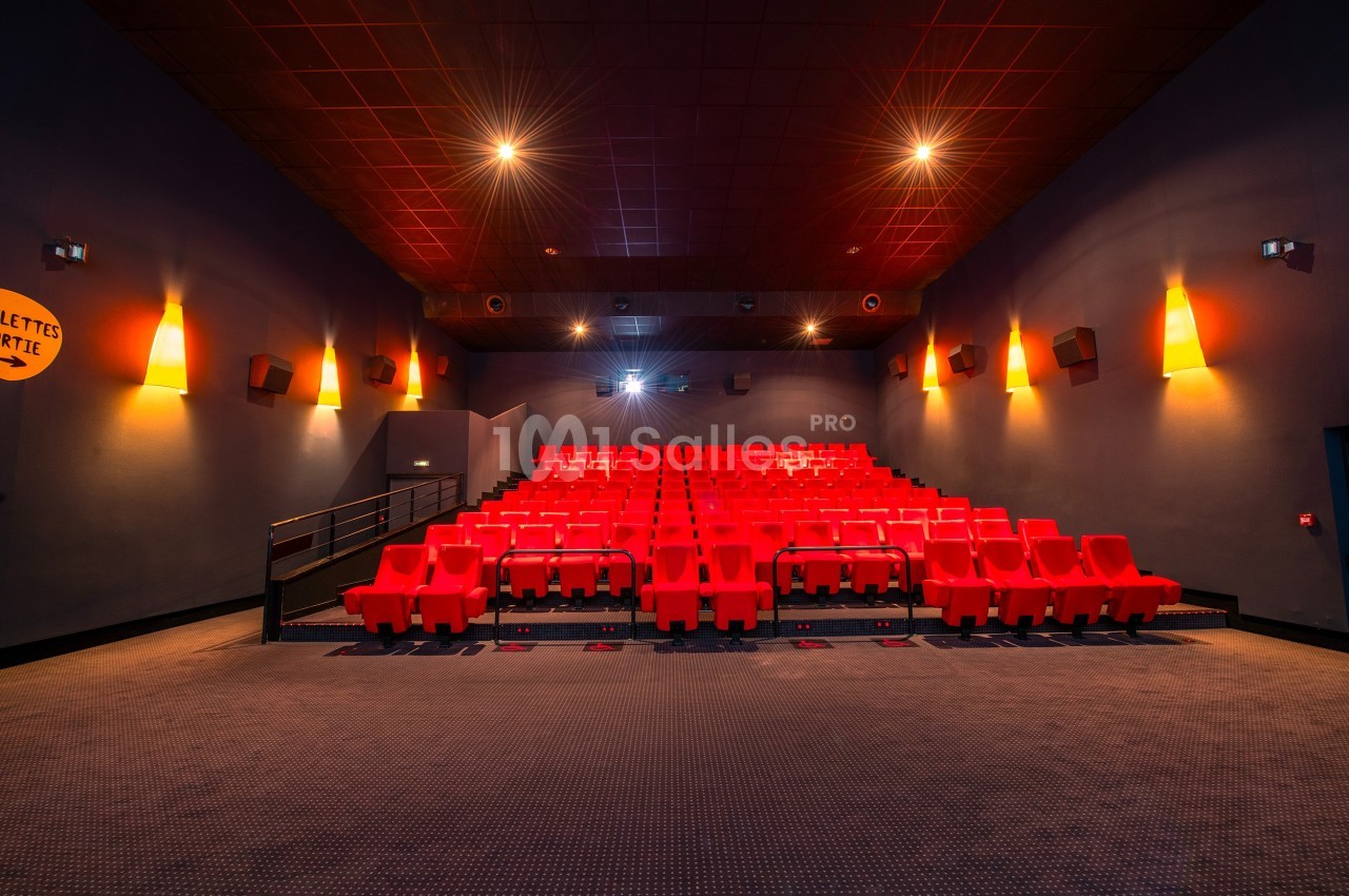 Salle de cinéma vide avec rangées de sièges rouges, éclairage tamisé et écran éteint en arrière-plan.