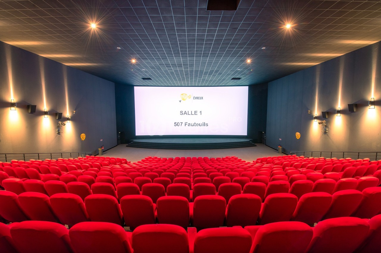 Salle de cinéma moderne avec des rangées de fauteuils rouges et un grand écran affichant ’Salle 1, 507 fauteuils’.