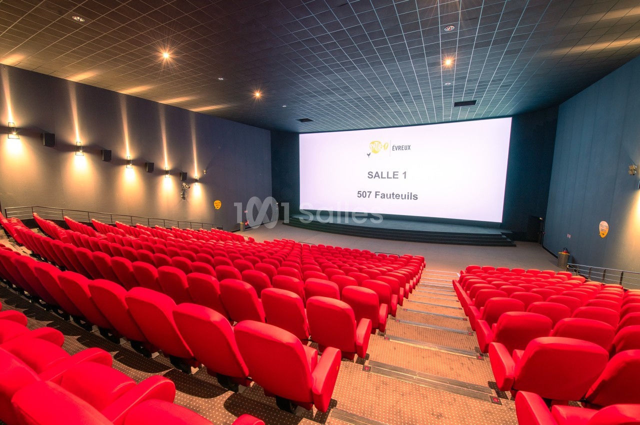 Salle de cinéma vide avec des rangées de fauteuils rouges, un grand écran blanc et un éclairage tamisé.