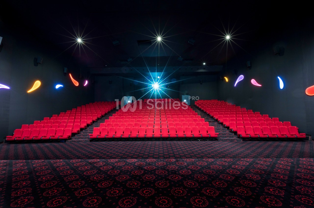 Salle de cinéma vide avec des rangées de sièges rouges, un projecteur allumé et un sol à motifs sombres.