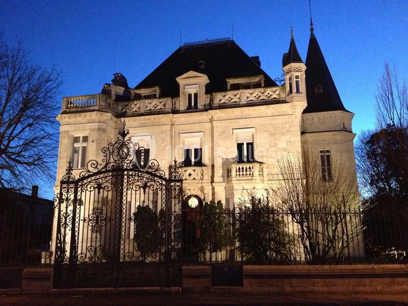 Façade d'un manoir éclairé en soirée, avec une grille en fer forgé au premier plan.