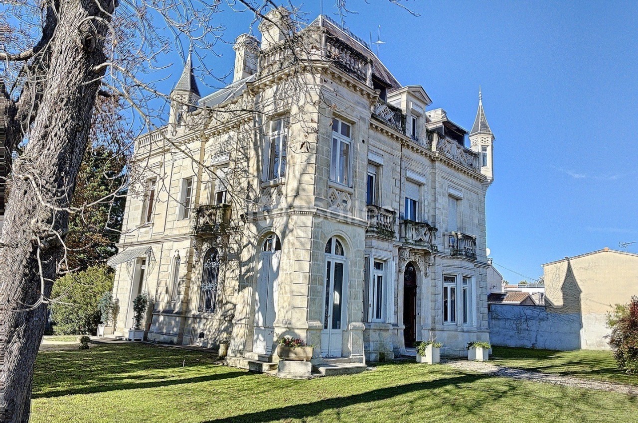 Façade d'un manoir en pierre avec tourelles, entouré d'un jardin arboré sous un ciel bleu.