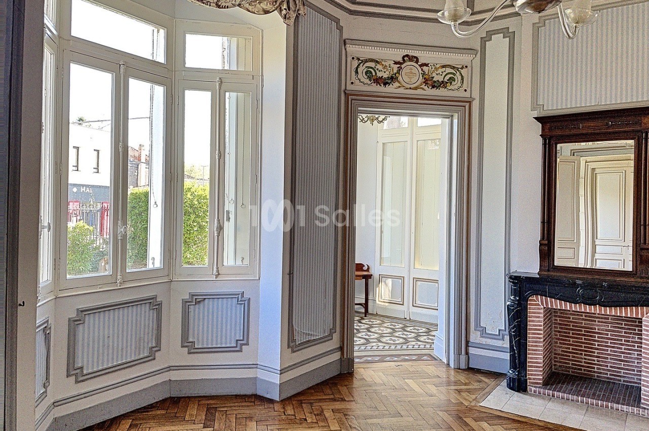 Intérieur lumineux avec parquet en bois, cheminée décorative, moulures au plafond et grandes fenêtres donnant sur l…