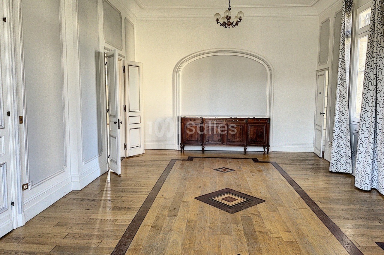 Salle vide avec parquet en bois clair, moulures blanches, buffet en bois et rideaux à motifs géométriques.