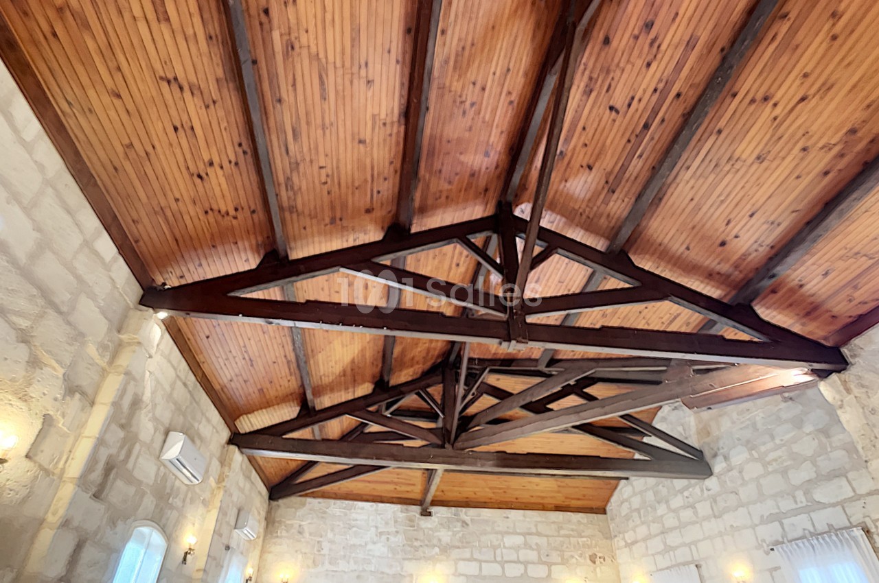 Plafond en bois avec poutres apparentes dans une salle aux murs en pierre, éclairée par des appliques murales.