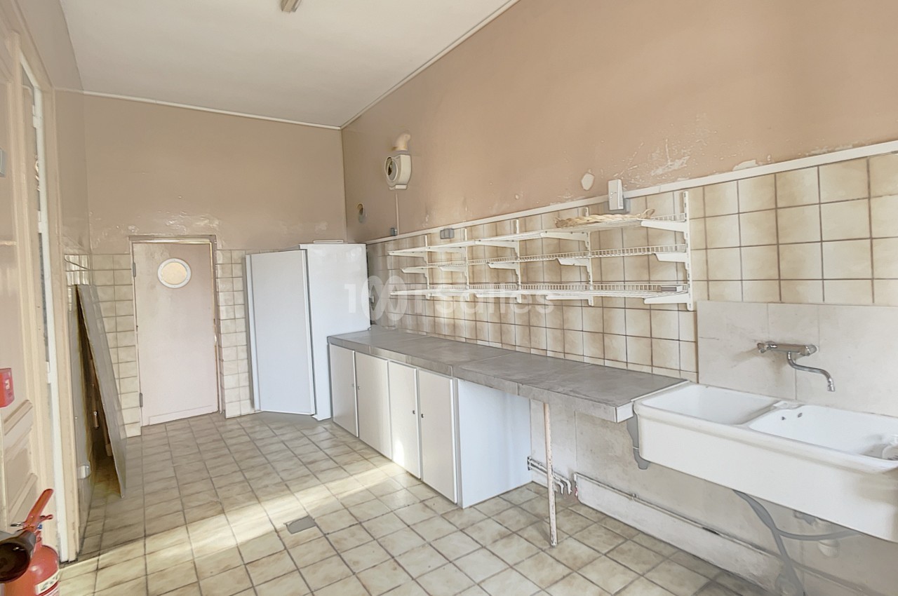 Cuisine vide avec carrelage beige, évier mural, étagères blanches, réfrigérateur et porte au fond.