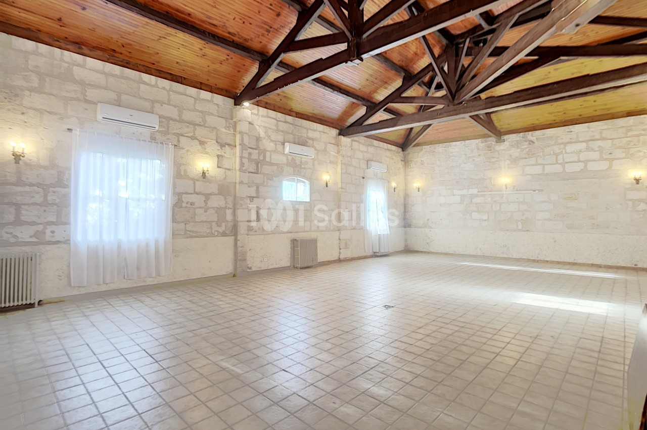 Salle spacieuse avec murs en pierre, plafond en bois à poutres apparentes et carrelage clair, éclairée par des fenêtres.