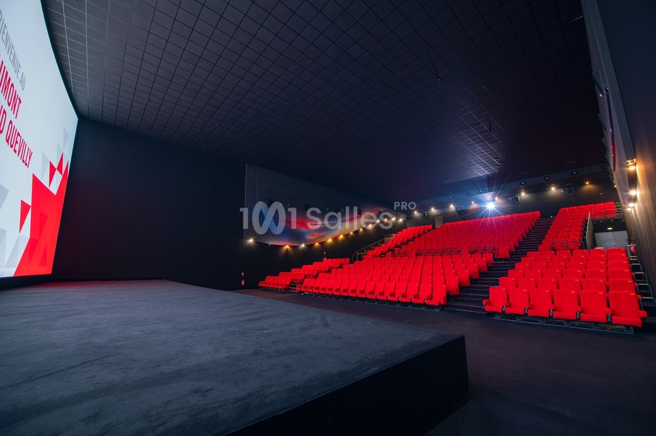 Salle de cinéma vide avec des rangées de sièges rouges, un écran blanc et un éclairage tamisé.