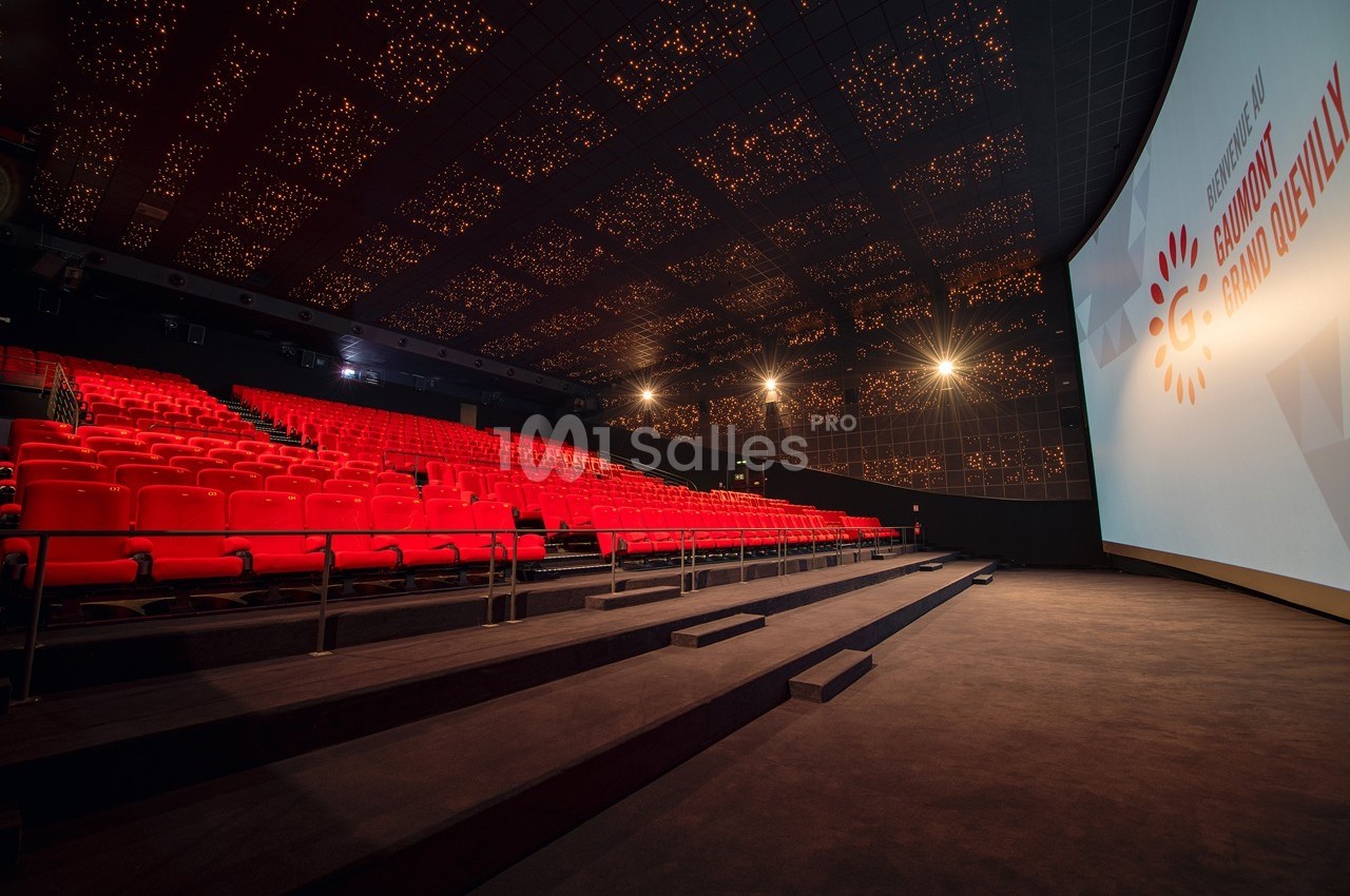 Salle de cinéma vide avec fauteuils rouges, grand écran allumé et plafond illuminé par des motifs lumineux.
