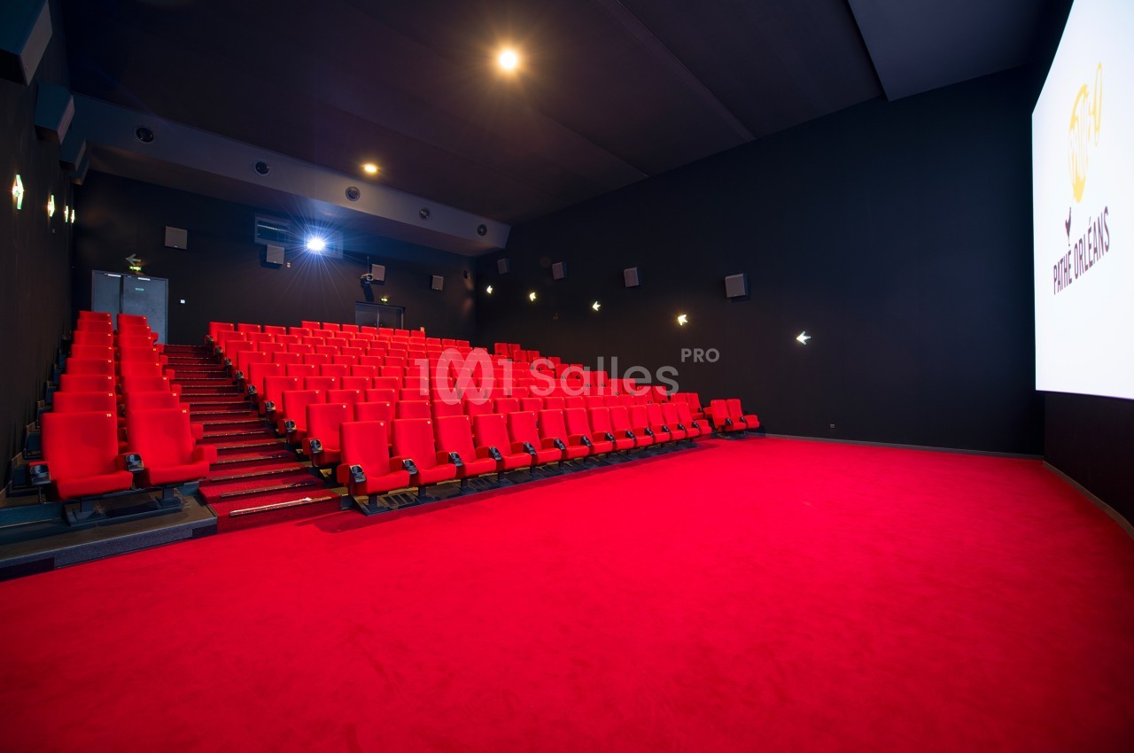 Salle de cinéma vide avec des rangées de fauteuils rouges, un écran blanc allumé et un sol recouvert de moquette rouge.