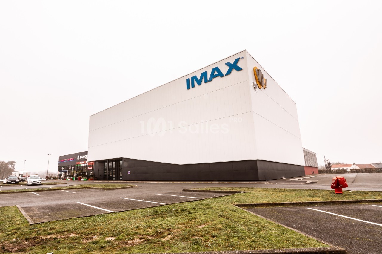 Façade d'un cinéma IMAX moderne avec parking vide au premier plan et ciel nuageux en arrière-plan.