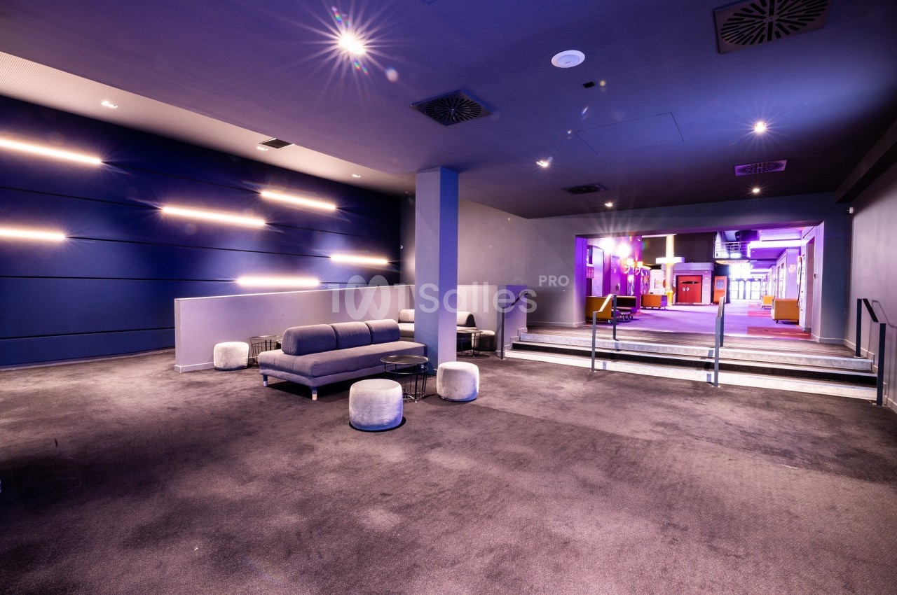 Espace intérieur moderne avec moquette, canapé gris, poufs et éclairage mural, menant à un couloir lumineux.
