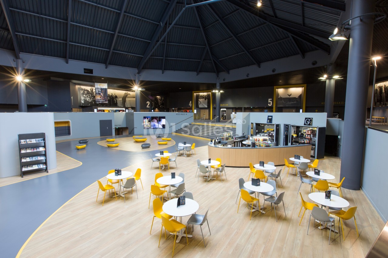 Salle spacieuse avec tables et chaises jaunes et grises, comptoir de café et numéros indiquant différentes sections.
