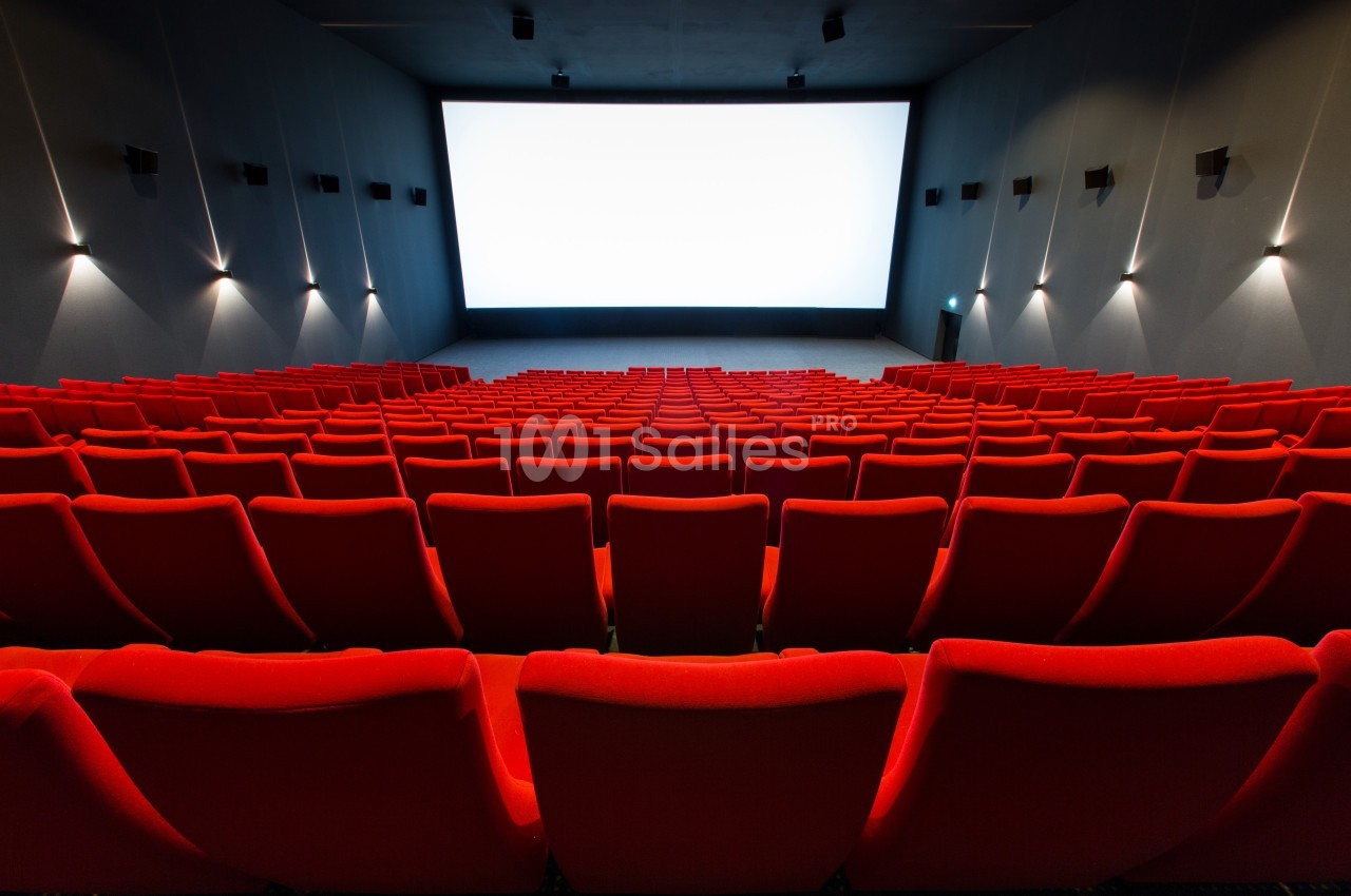 Salle de cinéma vide avec des rangées de fauteuils rouges et un grand écran blanc allumé à l'arrière-plan.