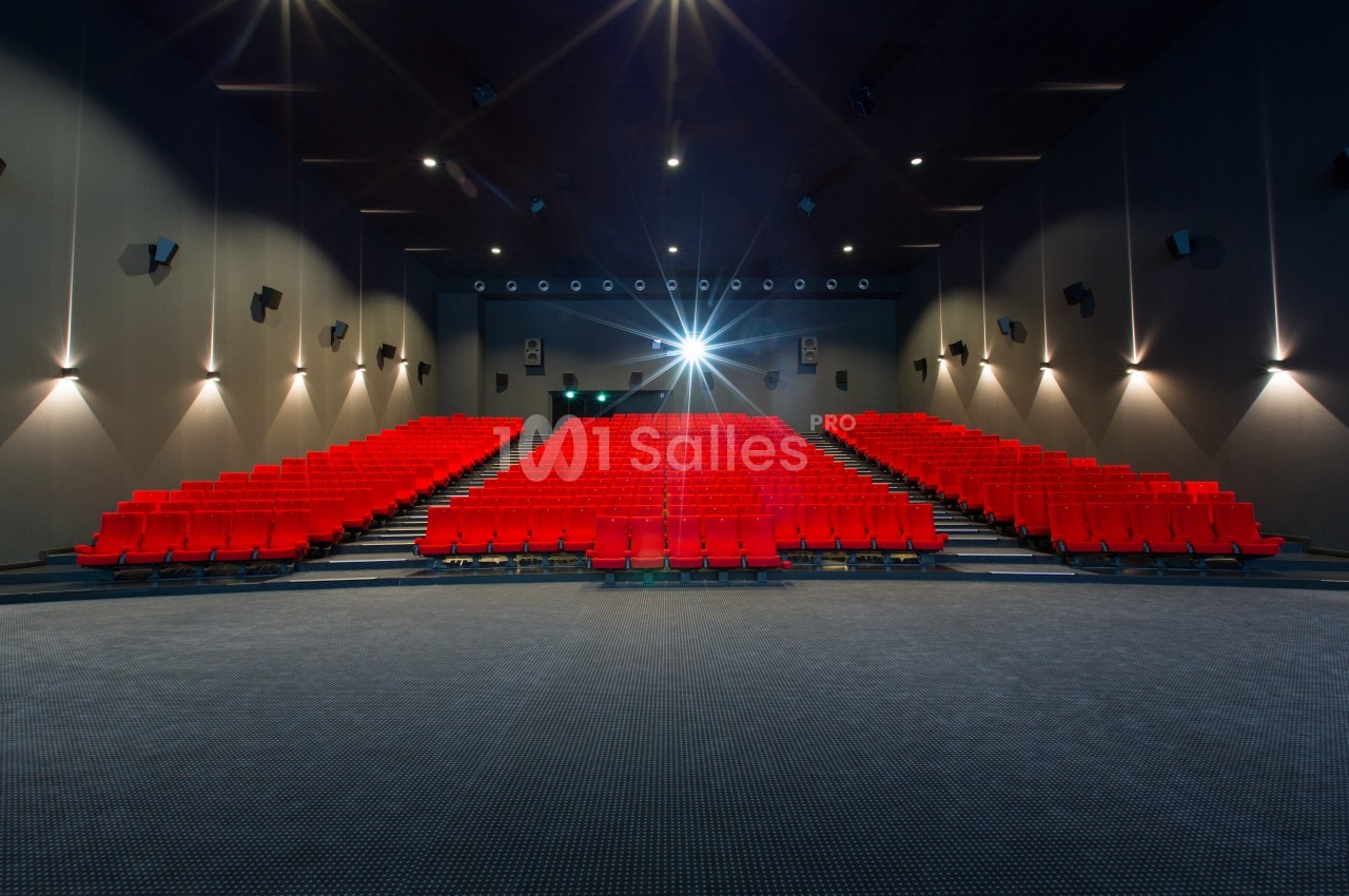 Salle de cinéma vide avec des rangées de sièges rouges et un projecteur allumé au fond.