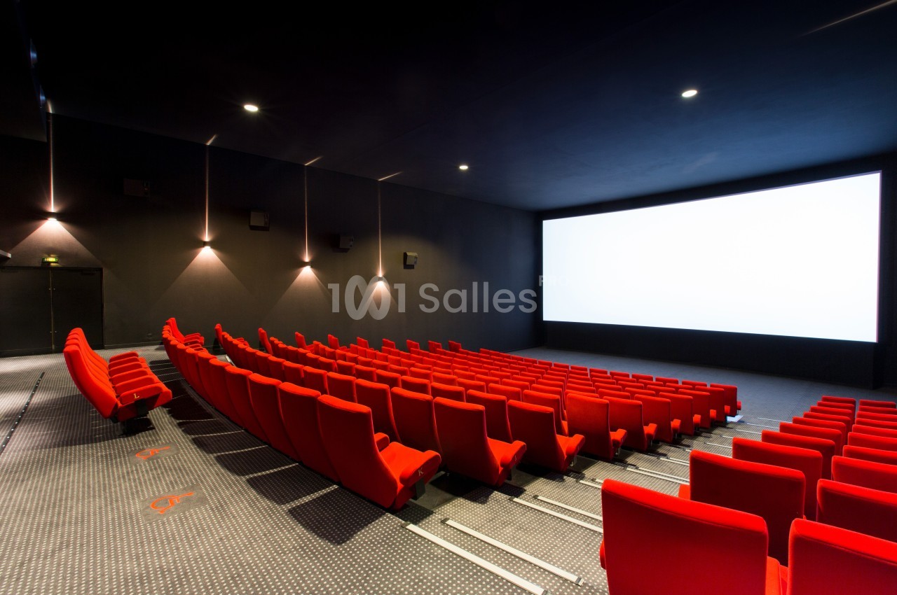 Salle de cinéma vide avec des rangées de sièges rouges et un grand écran blanc allumé.