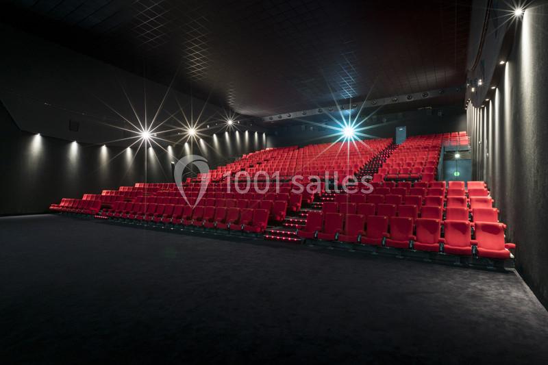 Salle de cinéma vide avec rangées de sièges rouges, éclairage tamisé et écran éteint en arrière-plan.