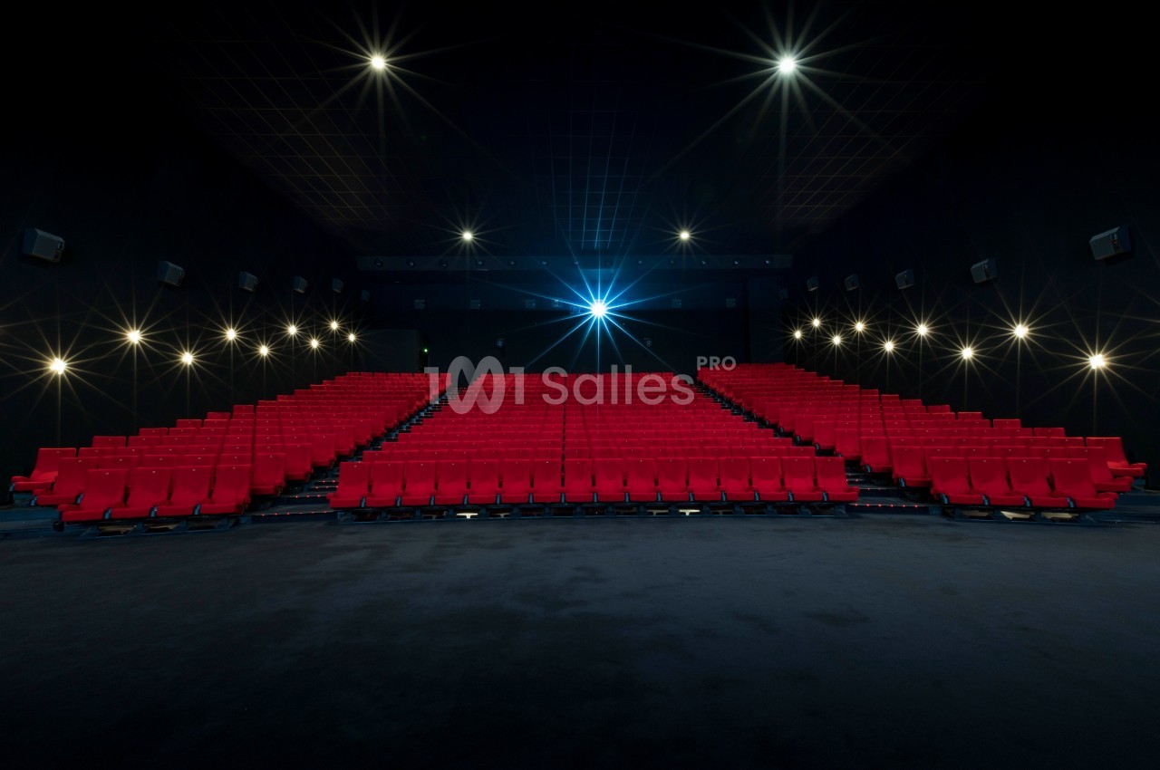 Salle de cinéma vide avec des rangées de sièges rouges et un écran allumé au fond, éclairée par des lumières tamisées.