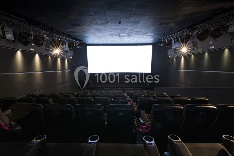 Salle de cinéma vide avec rangées de sièges noirs et un écran blanc allumé au fond.
