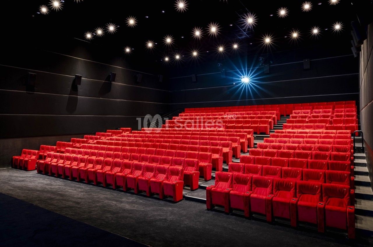 Salle de cinéma vide avec des rangées de fauteuils rouges et un projecteur allumé.