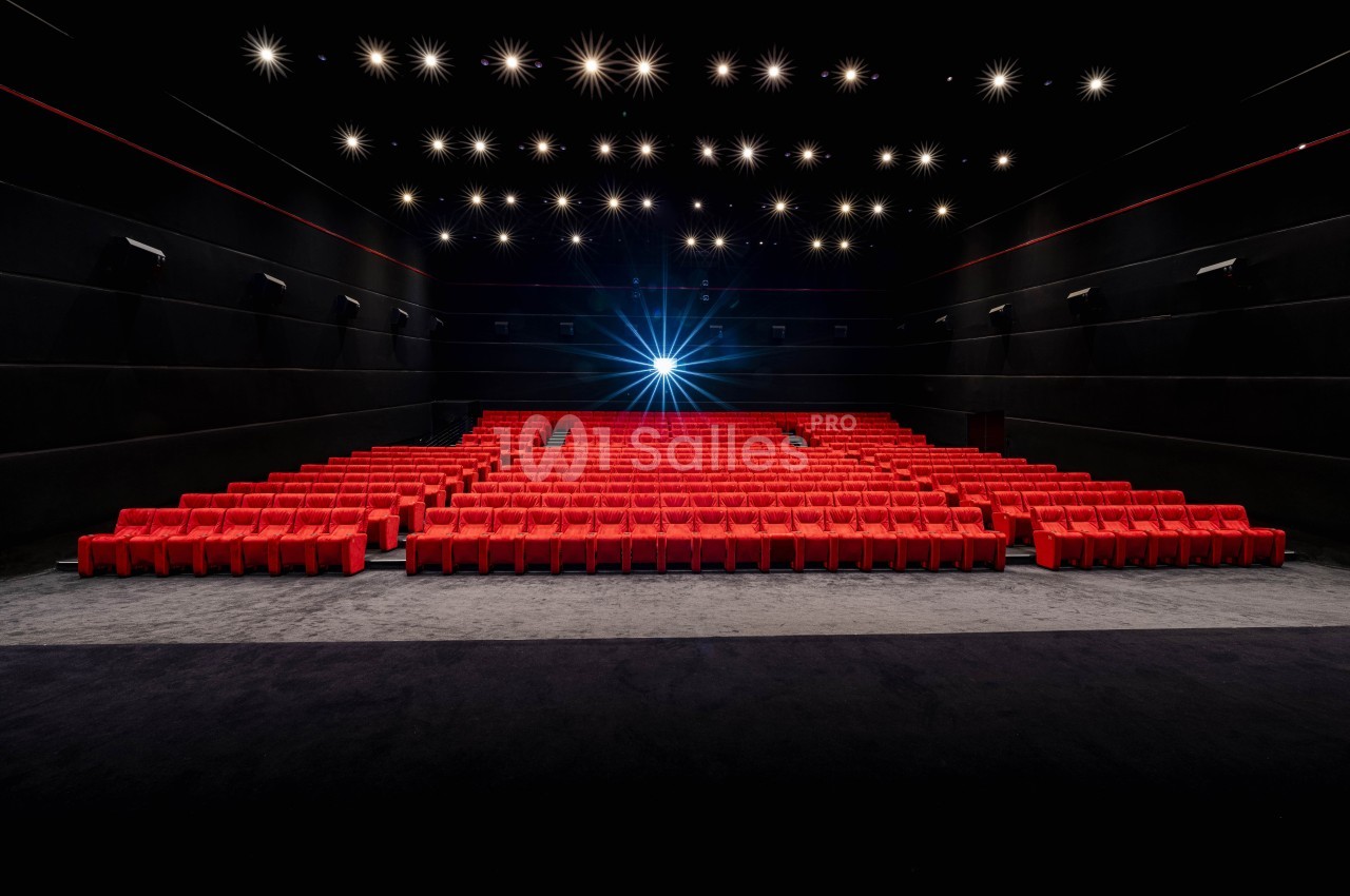 Salle de cinéma vide avec des rangées de sièges rouges et un projecteur allumé au fond.
