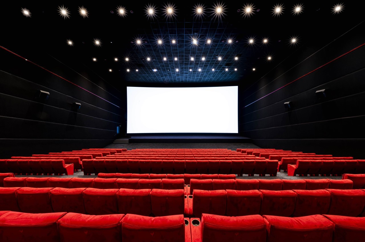 Salle de cinéma vide avec des rangées de fauteuils rouges et un écran blanc illuminé au fond.