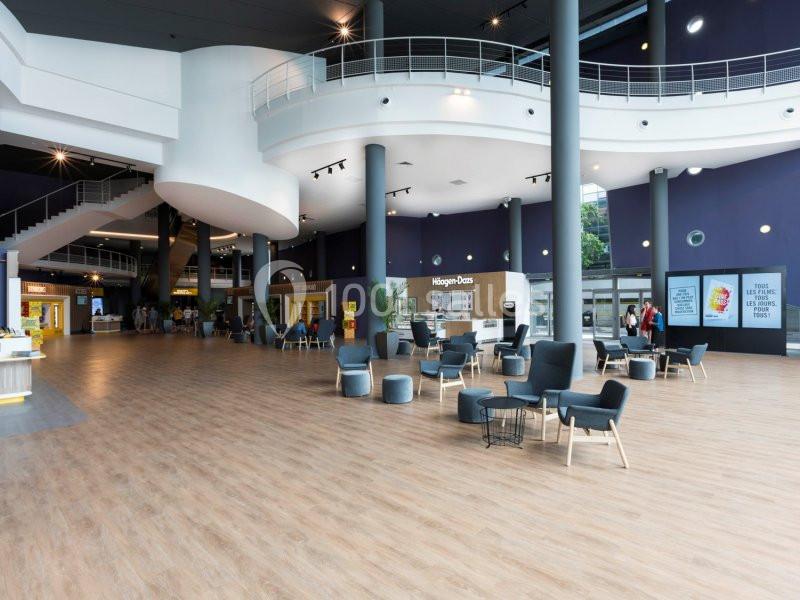 Espace intérieur spacieux avec sol en bois, fauteuils, tables, et éclairage naturel provenant de grandes fenêtres.